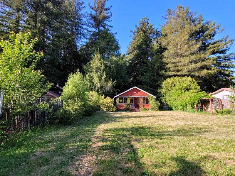 A small red cabin with a porch sits at the edge of a wide grassy yard in Del Norte County, surrounded by trees and bushes beneath a clear blue sky.
