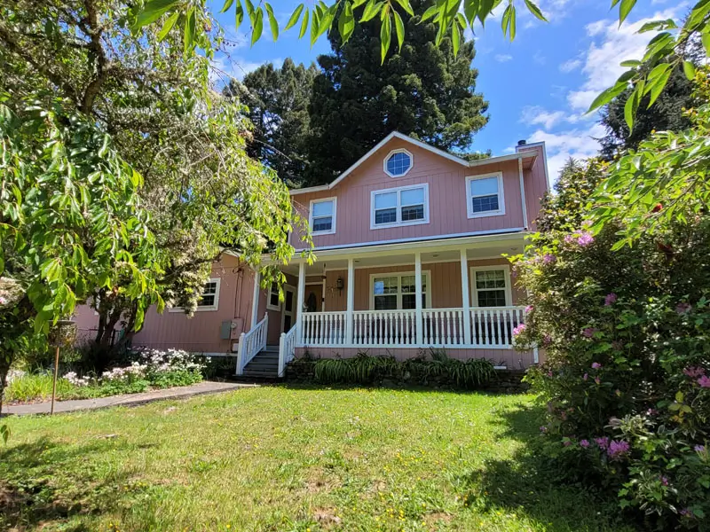 A pink two-story house with white trim and a front porch, surrounded by green trees, bushes, and a grassy lawn beneath a partly cloudy blue sky — reflecting classic Humboldt charm.
